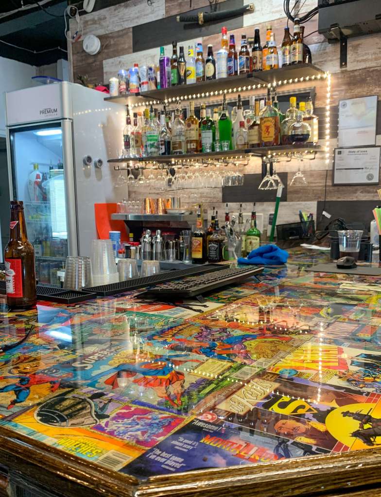 Picture of a bartop featuring a collage of comics and bottle of liquor and alcholo behind the bar